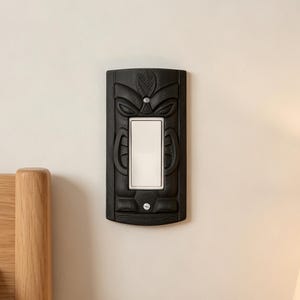 May include: A black rectangular light switch cover plate featuring a carved tiki-inspired face design. The plate surrounds a white rectangular rocker light switch and is secured to the wall with two visible silver-colored metal screws.