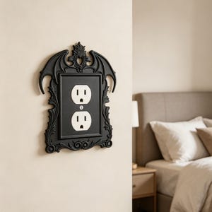 May include: A black wall plate cover for a duplex electrical outlet, featuring an ornate frame with decorative bat wing motifs at the top and intricate scrollwork along the sides, surrounding a standard white two-socket electrical receptacle.