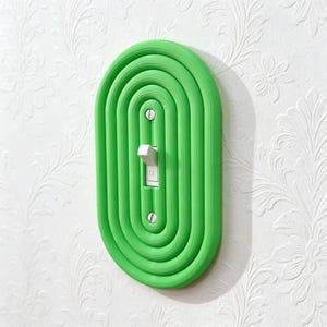 Può includere: Una copertura per interruttore della luce verde brillante con un design ovale a strati. La copertura circonda un interruttore della luce standard con un interruttore a levetta bianco. La copertura è montata su una parete bianca con un motivo floreale.