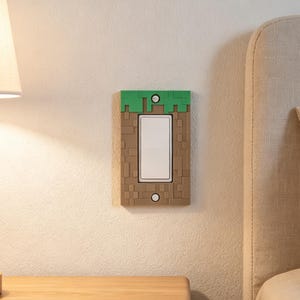 May include: A rectangular light switch cover plate featuring a pixelated design with a green top section and a brown block-patterned lower section. The cover is secured with two silver screws and surrounds a white vertical rocker light switch.