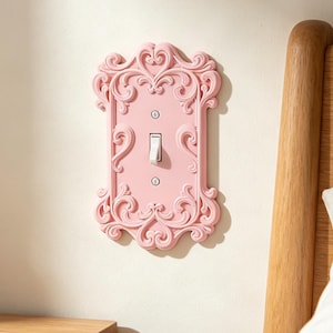 May include: A light pink light switch cover plate featuring an ornate, raised scrollwork border with symmetrical swirling patterns. The rectangular plate includes a central vertical toggle switch and two small silver screws for mounting.