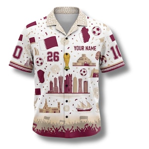 Peut inclure: Chemise blanche &agrave; motifs bordeaux et beige. La chemise pr&eacute;sente un ballon de football, le drapeau du Qatar et le texte "YOUR NAME". La chemise porte les num&eacute;ros 0, 10 et 26. La chemise a un col et des manches courtes.