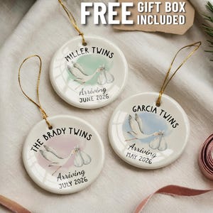 Può includere: Tre ornamenti rotondi in ceramica bianca con cordini dorati. Ogni ornamento presenta una cicogna che trasporta un bambino, le parole "Twins" e un cognome. Gli ornamenti includono anche il testo "Arriving" e un mese e un anno.