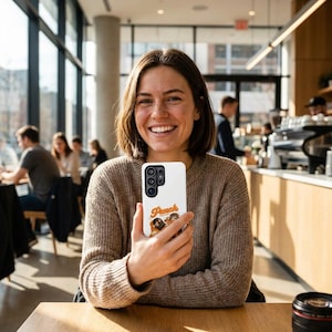 Puede incluir: Una funda blanca para smartphone con la palabra "Peach" en escritura naranja, con dos personajes de dibujos animados. La funda la sostiene una persona con un suéter marrón, sentada en una mesa en un café.