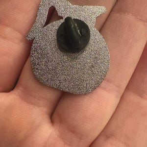 May include: A silver-colored enamel pin in the shape of a teapot, with a black pin back. The pin has a textured surface and a small cutout near the top. The pin is shown against a neutral background.
