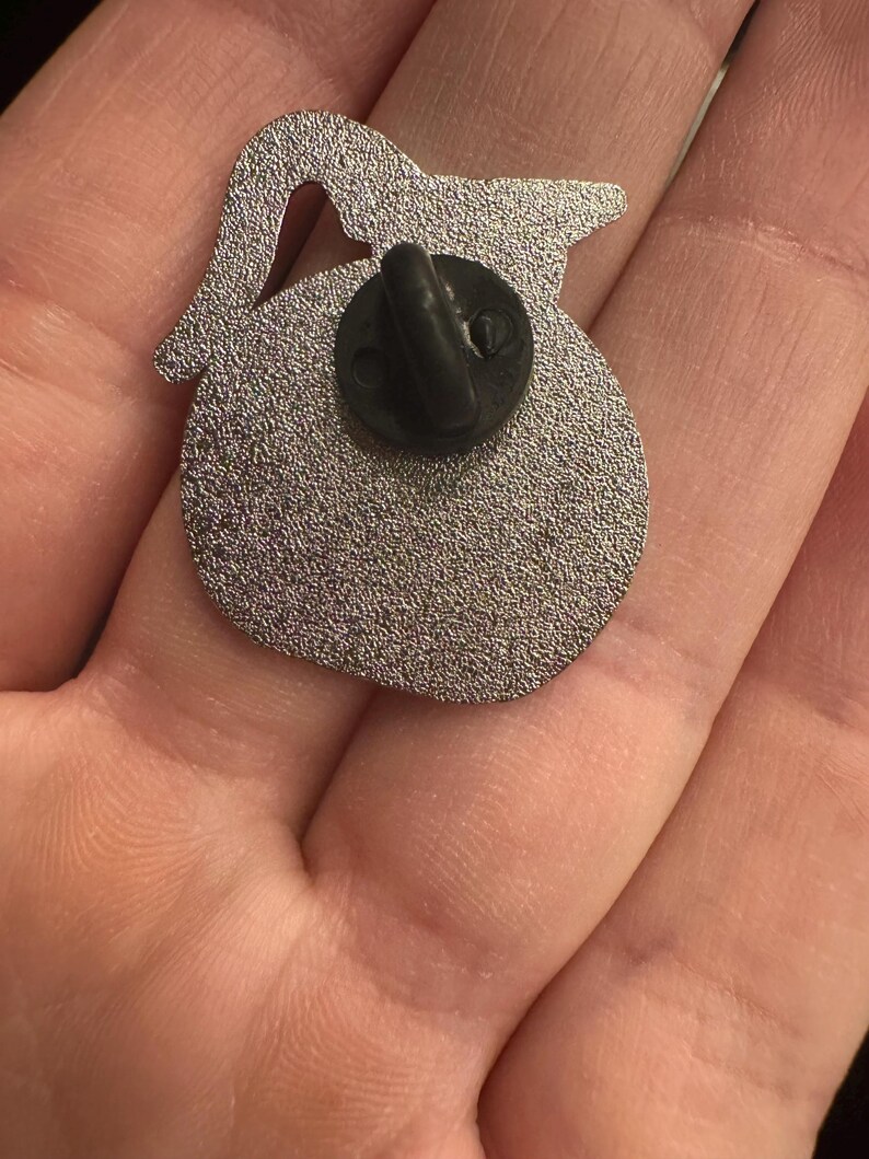 May include: A silver-colored enamel pin in the shape of a teapot, with a black pin back. The pin has a textured surface and a small cutout near the top. The pin is shown against a neutral background.