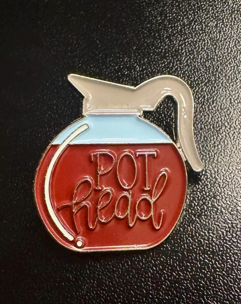 May include: A red and blue enamel pin in the shape of a coffee pot. The pin features the words "POT head" in a cursive font. The pin has a silver-colored handle and spout, and a white outline.