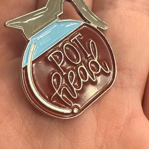 May include: A pin shaped like a coffee pot with the words "POT head" in white script. The pin has a brown body, a light blue top, and a silver handle and trim. A novelty item.