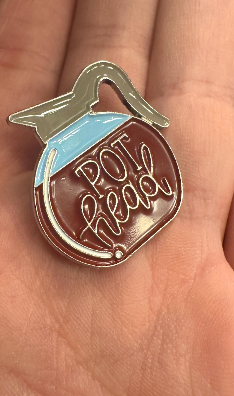 May include: A pin shaped like a coffee pot with the words "POT head" in white script. The pin has a brown body, a light blue top, and a silver handle and trim. A novelty item.