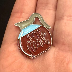 May include: Enamel pin in the shape of a coffee pot with the words "POT head" in cursive. The pin has a brown body, a light blue top, and a silver handle and trim. The pin is held in a person's hand.