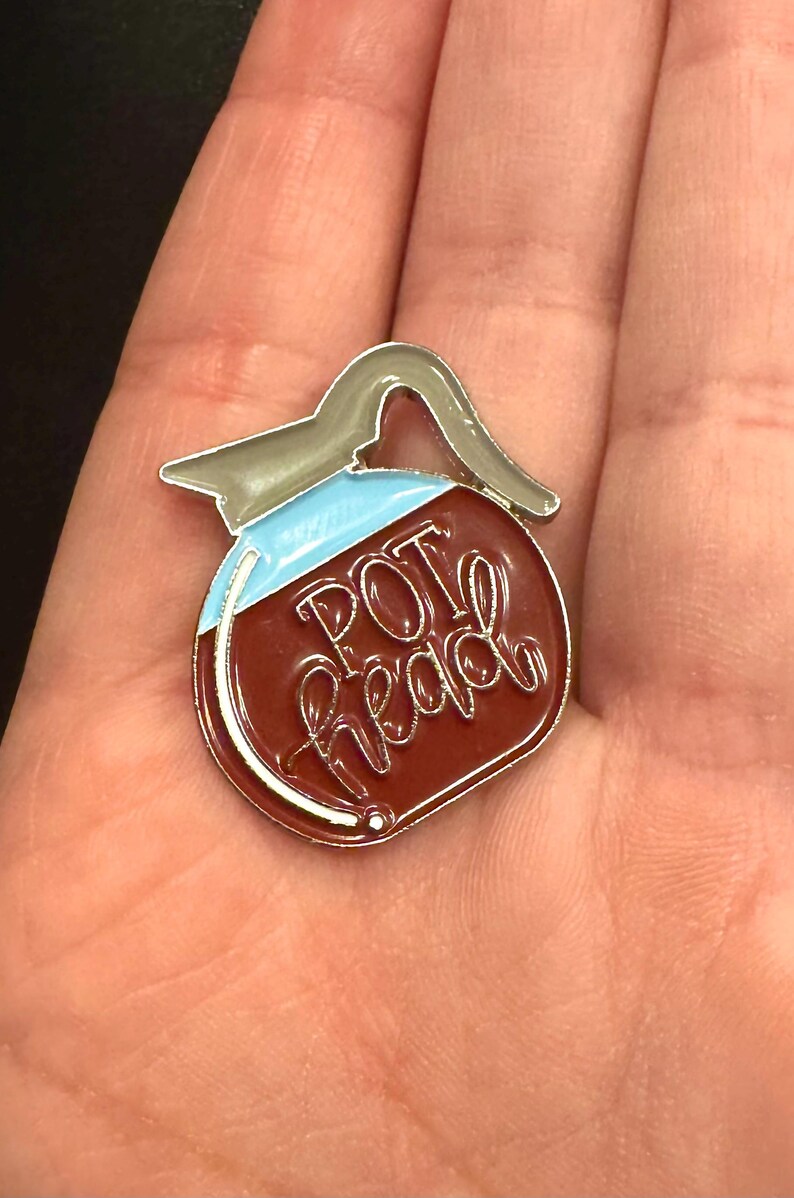 May include: Enamel pin in the shape of a coffee pot with the words "POT head" in cursive. The pin has a brown body, a light blue top, and a silver handle and trim. The pin is held in a person's hand.