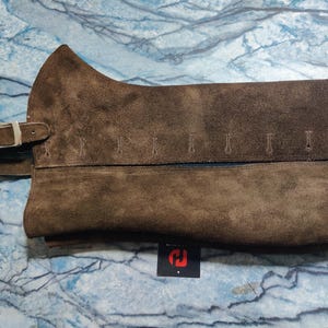 Può includere: Chaps in pelle scamosciata marrone con chiusura a fibbia e cuciture decorative. Gli chaps sono di un ricco colore marrone e sembrano essere realizzati in un materiale morbido e strutturato. Il design include una fibbia in stile cintura e una serie di piccole cuciture verticali.