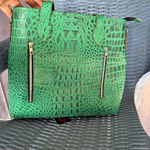 Puede incluir: Bolso tote verde con textura de cocodrilo, dos cremalleras frontales y un asa superior. El bolso tiene forma rectangular y un interior marrón. El bolso está sobre una superficie tejida azul.
