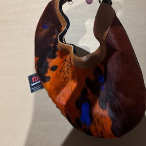 May include: A small, crescent-shaped handbag with a unique spotted pattern in shades of brown, orange, and black. The bag features a tan leather trim and a gold-colored metal handle. Blue paint splatters add a pop of color to the design.
