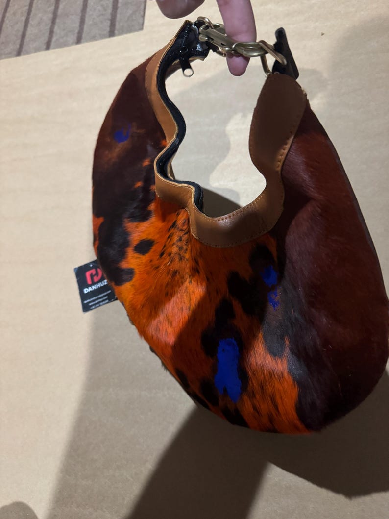 May include: A small, crescent-shaped handbag with a unique spotted pattern in shades of brown, orange, and black. The bag features a tan leather trim and a gold-colored metal handle. Blue paint splatters add a pop of color to the design.
