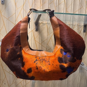 May include: A brown and orange cowhide shoulder bag with a leather strap. The bag has a unique pattern with dark brown and orange fur, and blue accents. The strap is a lighter brown leather.