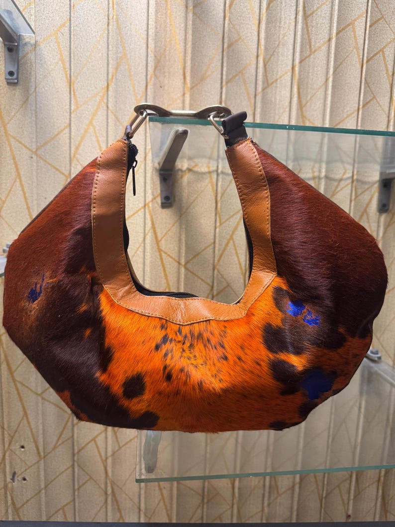 May include: A brown and orange cowhide shoulder bag with a leather strap. The bag has a unique pattern with dark brown and orange fur, and blue accents. The strap is a lighter brown leather.