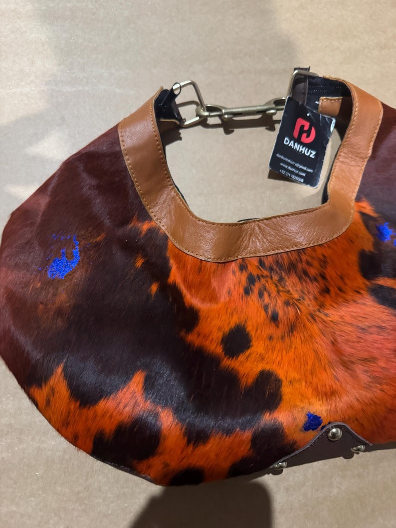 May include: A shoulder bag with a unique pattern of brown, orange, and black fur. The bag has a brown leather trim and a metal chain strap. A tag with the brand name "DANHUZ" is attached.