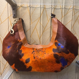 May include: A brown and orange cowhide handbag with a leather handle and strap. The bag has a unique pattern with dark brown and orange fur, and blue paint splatters. A gold-colored clasp is attached to the handle.