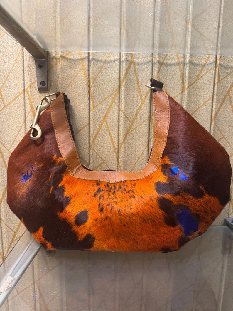 May include: A brown and orange cowhide handbag with a leather handle and strap. The bag has a unique pattern with dark brown and orange fur, and blue paint splatters. A gold-colored clasp is attached to the handle.
