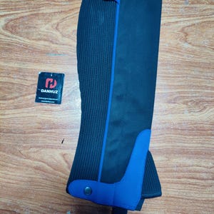 Peut inclure: Guêtres d'équitation noires et bleues avec un panneau côtelé vertical et un accent bleu. Les guêtres ont une fermeture à bouton-pression et une sangle en bas. L'étiquette de la marque "DANHUZ" est visible.