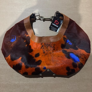 May include: A brown, orange, and black patterned handbag with a leather strap and accents. The bag has a unique spotted design with blue accents and a metal chain. The tag reads DANHUZ.