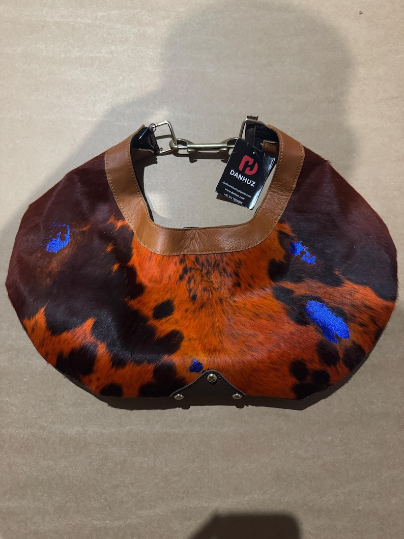 May include: A brown, orange, and black patterned handbag with a leather strap and accents. The bag has a unique spotted design with blue accents and a metal chain. The tag reads DANHUZ.