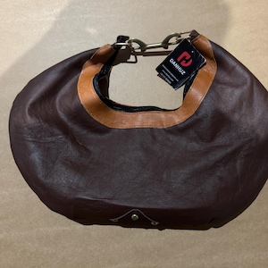 May include: A brown leather hobo bag with a tan trim around the opening. The bag has a curved shape, a zipper closure, and a metal handle. A tag with the brand name "DANHUZ" is attached.
