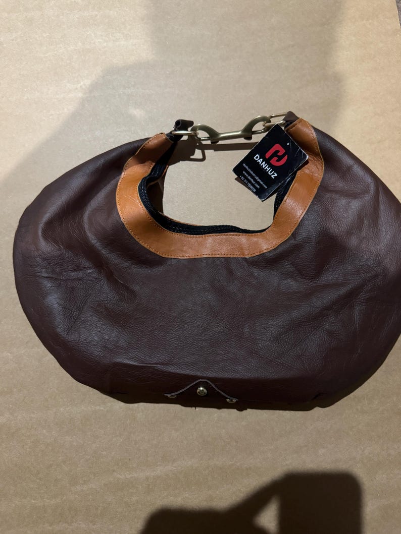 May include: A brown leather hobo bag with a tan trim around the opening. The bag has a curved shape, a zipper closure, and a metal handle. A tag with the brand name "DANHUZ" is attached.
