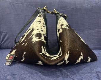 May include: A crescent-shaped shoulder bag featuring a brown and white cowhide pattern with black leather trim, gold-tone metal hardware, and a black strap. A small rectangular tag with the text DANHUI is attached to the side of the bag.
