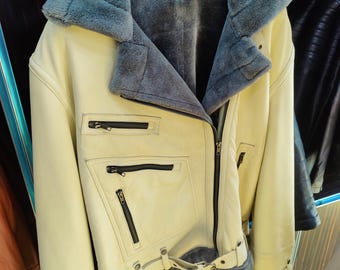 May include: Cream-colored leather jacket with a gray faux fur collar and lining. The jacket features black zippers, a belt, and a classic biker style. The jacket is hanging on a black hanger.
