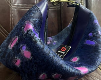 May include: A crescent-shaped handbag crafted from dark blue and black animal hide with vibrant pink patches. The bag features a smooth dark blue leather trim, a top zipper closure, a brass-toned metal clasp, and a black tag labeled DANHUZ.