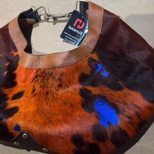 May include: A unique, handcrafted collar made from animal hide. The collar features a brown leather trim and a vibrant pattern of orange, black, and blue accents. The tag reads "DANHUZ".