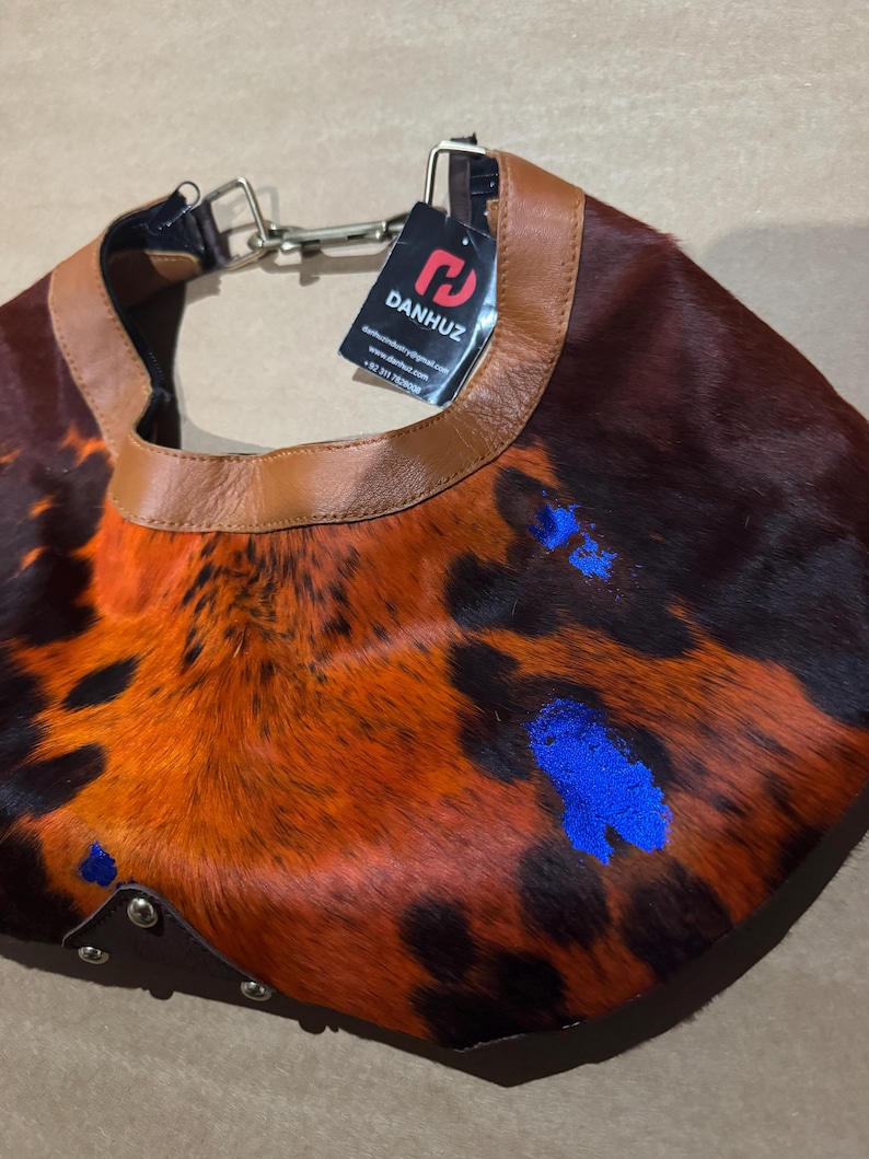 May include: A unique, handcrafted collar made from animal hide. The collar features a brown leather trim and a vibrant pattern of orange, black, and blue accents. The tag reads "DANHUZ".