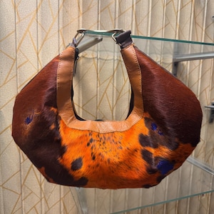 May include: A brown and orange patterned handbag with a leather handle. The bag has a unique design with a mix of brown and orange fur, accented with dark blue spots. The handle is made of brown leather and is attached to the bag with metal hardware.