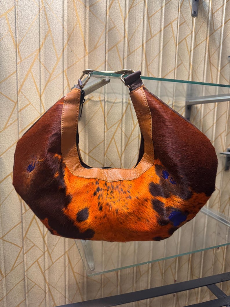 May include: A brown and orange patterned handbag with a leather handle. The bag has a unique design with a mix of brown and orange fur, accented with dark blue spots. The handle is made of brown leather and is attached to the bag with metal hardware.