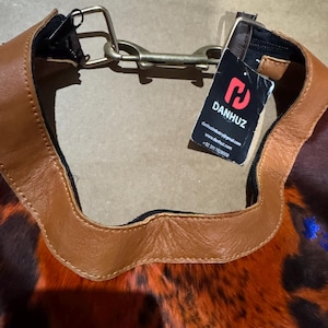 May include: A close-up of a handbag with a brown leather trim and a patterned body in shades of orange, brown, and black. The bag has a metal clasp and a tag with the brand name "DANHUZ".