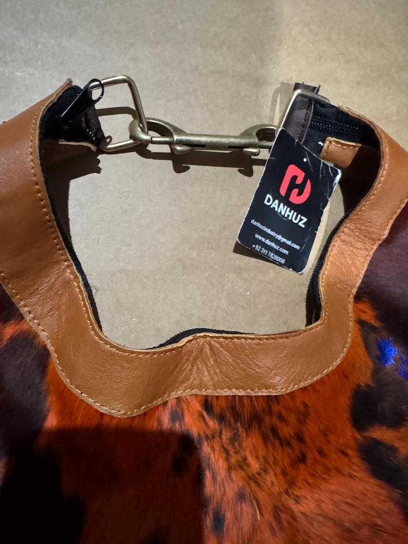 May include: A close-up of a handbag with a brown leather trim and a patterned body in shades of orange, brown, and black. The bag has a metal clasp and a tag with the brand name "DANHUZ".