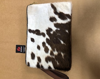 May include: A rectangular clutch bag with a cowhide pattern in white and brown. The bag has a gold zip and a brown leather wrist strap. A black tag with red text is attached to the side.
