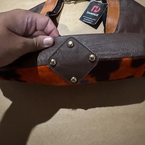 May include: A brown leather handbag with orange and black patterned accents. The bag features gold-colored hardware, including a clasp and decorative studs. A tag with the brand name "DANHUZ" is attached.