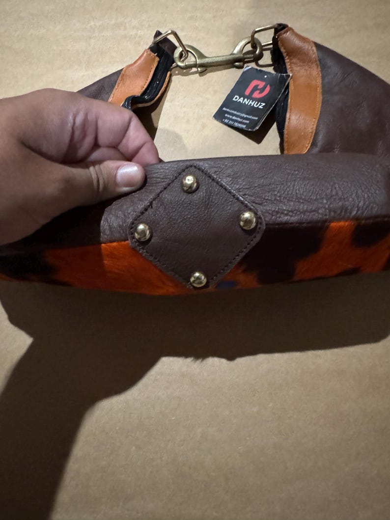 May include: A brown leather handbag with orange and black patterned accents. The bag features gold-colored hardware, including a clasp and decorative studs. A tag with the brand name "DANHUZ" is attached.