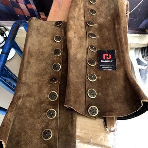 May include: Brown suede leather chaps with brass buttons. The chaps have a classic design, with multiple buttons down the side and a flared bottom. The product tag reads "DANHUZ".