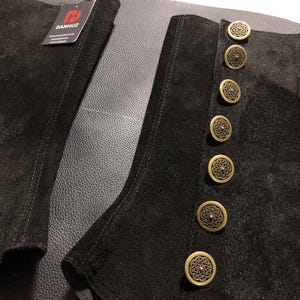 May include: Close-up of black suede boot cuffs with decorative gold-colored buttons. The cuffs have a tailored design with visible stitching. The buttons are round with an ornate, openwork pattern. The brand tag is visible.