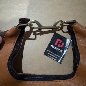 May include: Close-up of a brown and tan leather bag with a black zipper and a brass-colored clasp. A tag with the brand name "DANHUZ" is attached. The bag appears to be new and unused.