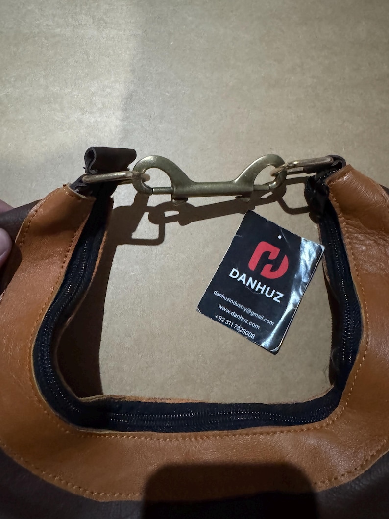 May include: Close-up of a brown and tan leather bag with a black zipper and a brass-colored clasp. A tag with the brand name "DANHUZ" is attached. The bag appears to be new and unused.