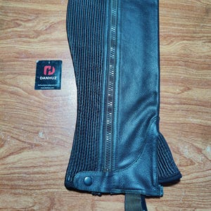 May include: Black leather riding chaps with a full-length zipper and a ribbed panel on the side. The chaps have a button closure at the bottom and an adjustable strap. The brand tag "DANHUZ" is visible.