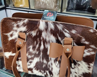 May include: A brown and white cowhide handbag with brown leather handles and accents. The bag has a rectangular shape with a top zipper closure. The brand name "DANHUZ" is visible on a tag attached to the bag.