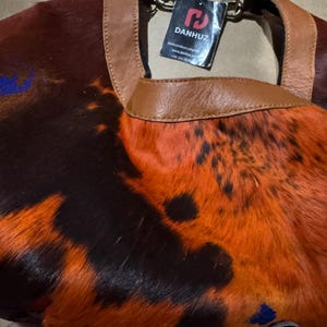 May include: A handbag with a unique pattern of orange, brown, and black fur. The bag has a brown leather handle and trim. A tag with the brand name "DANHUZ" is attached to the handle.