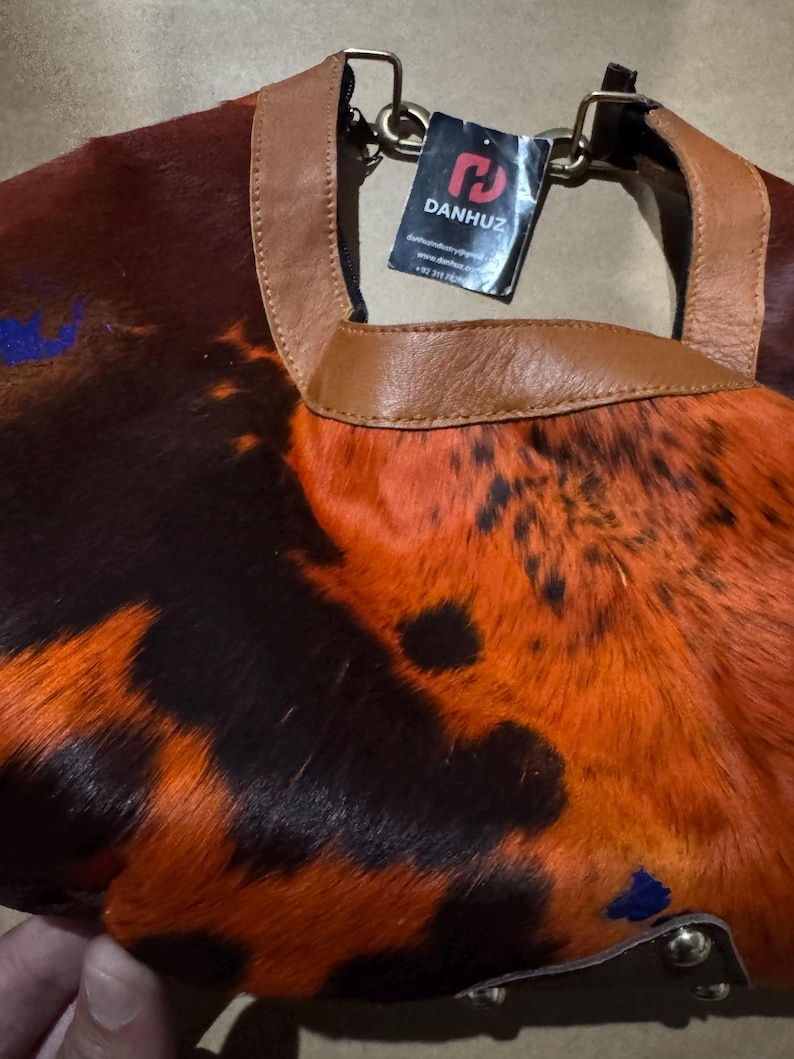 May include: A handbag with a unique pattern of orange, brown, and black fur. The bag has a brown leather handle and trim. A tag with the brand name "DANHUZ" is attached to the handle.