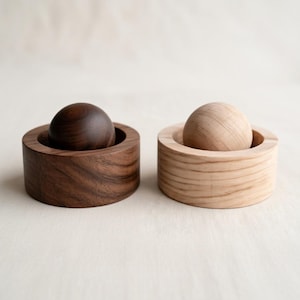May include: Two wooden decorative objects. One is made of dark walnut, and the other is made of light wood. Each has a spherical top set in a circular base. The dark wood object has a rich, warm tone, while the light wood object has a natural, pale color.